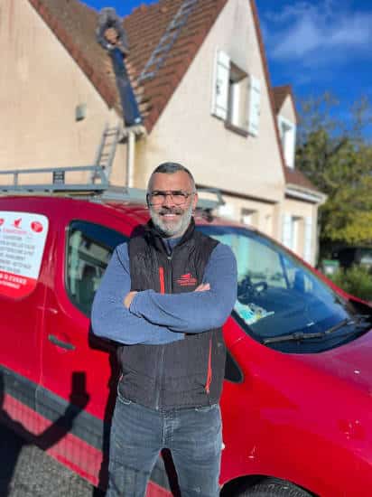 Portrait de Monsieur Moïse Fortin près de son véhicule avec en arrière-plan une maison en cours de réparation à Gazeran l'artisan est en train d'intervenir sur une toiture.